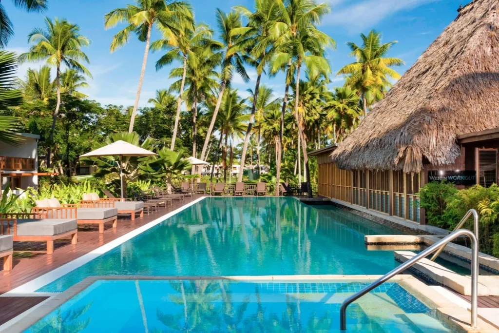 Outdoor swimming pool at a tropical resort with lounge chairs, umbrellas, and a thatched-roof building surrounded by tall palm trees.